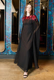 Elegant black maxi cape dress with a relaxed, fluid silhouette and a sheer flowing cape overlay that offers arm and shoulder coverage. The cape is decorated with deep red hand-sequinned floral motifs and cascading beaded tassels that add movement and spar