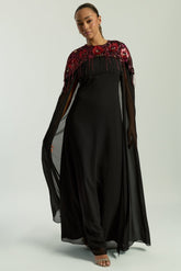 Elegant black maxi cape dress with a relaxed, fluid silhouette and a sheer flowing cape overlay that offers arm and shoulder coverage. The cape is decorated with deep red hand-sequinned floral motifs and cascading beaded tassels that add movement and spar
