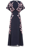 Navy floral embroidered maxi dress with pink detailing, worn by a model. Features flutter sleeves, a fitted waist and a flowing full-length skirt—perfect for weddings, evening occasions and special events.