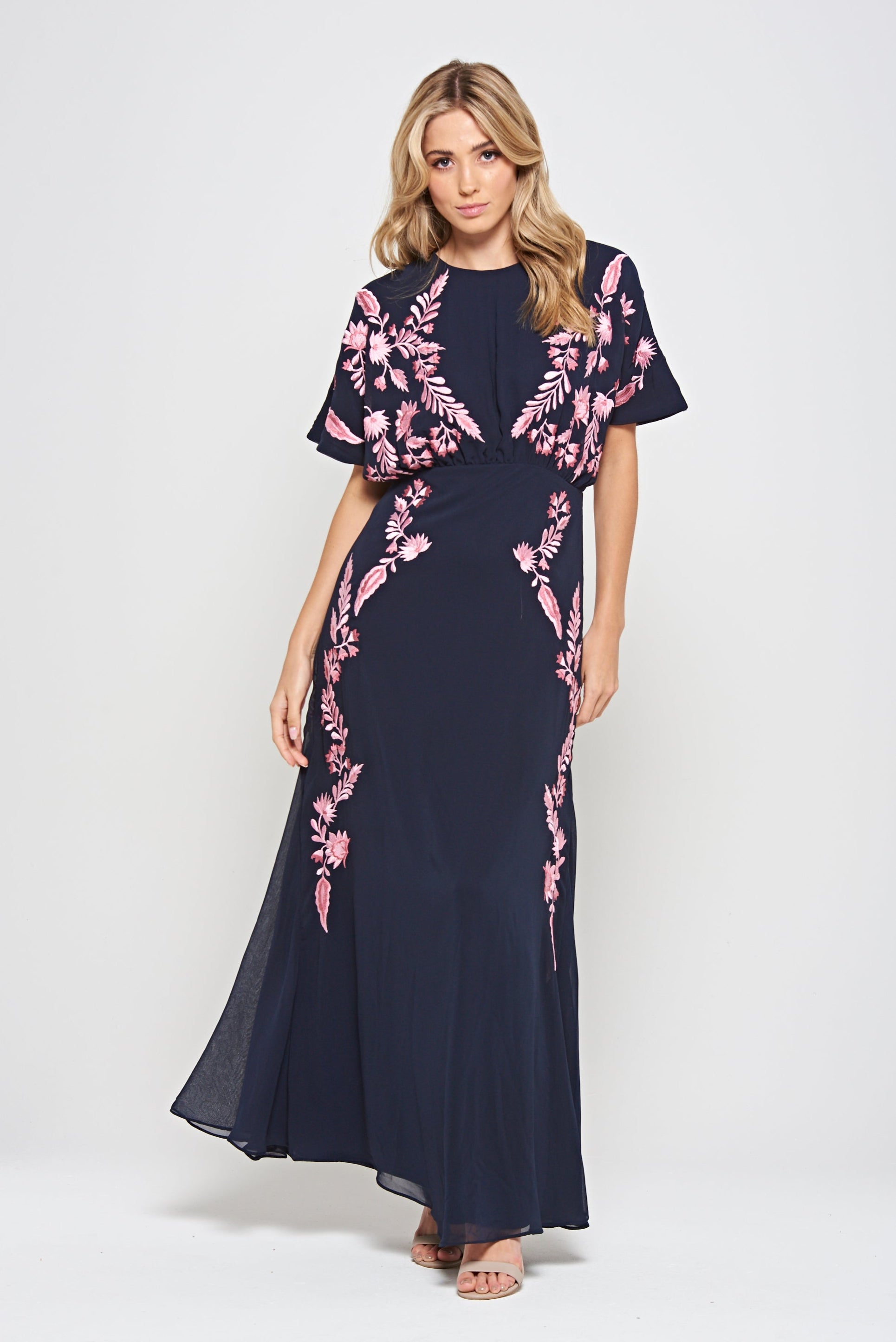 Navy floral embroidered maxi dress with pink detailing, worn by a model. Features flutter sleeves, a fitted waist and a flowing full-length skirt—perfect for weddings, evening occasions and special events.