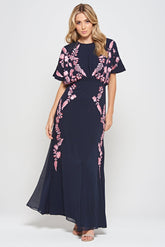 Navy floral embroidered maxi dress with pink detailing, worn by a model. Features flutter sleeves, a fitted waist and a flowing full-length skirt—perfect for weddings, evening occasions and special events.