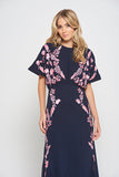 Navy floral embroidered maxi dress with pink detailing, worn by a model. Features flutter sleeves, a fitted waist and a flowing full-length skirt—perfect for weddings, evening occasions and special events.