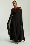 Elegant black maxi cape dress with a relaxed, fluid silhouette and a sheer flowing cape overlay that offers arm and shoulder coverage. The cape is decorated with deep red hand-sequinned floral motifs and cascading beaded tassels that add movement and spar