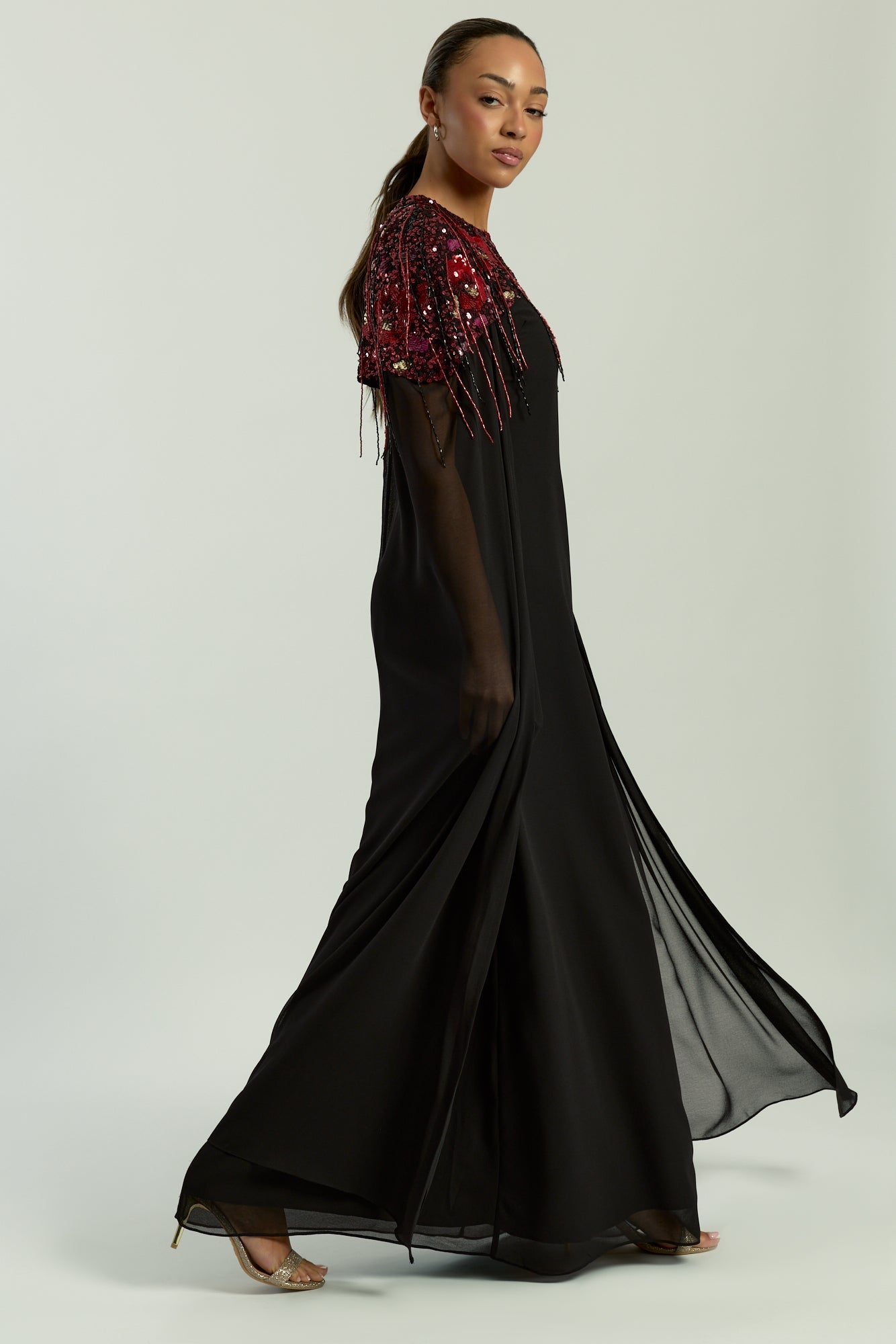 Elegant black maxi cape dress with a relaxed, fluid silhouette and a sheer flowing cape overlay that offers arm and shoulder coverage. The cape is decorated with deep red hand-sequinned floral motifs and cascading beaded tassels that add movement and spar