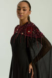 Elegant black maxi cape dress with a relaxed, fluid silhouette and a sheer flowing cape overlay that offers arm and shoulder coverage. The cape is decorated with deep red hand-sequinned floral motifs and cascading beaded tassels that add movement and spar