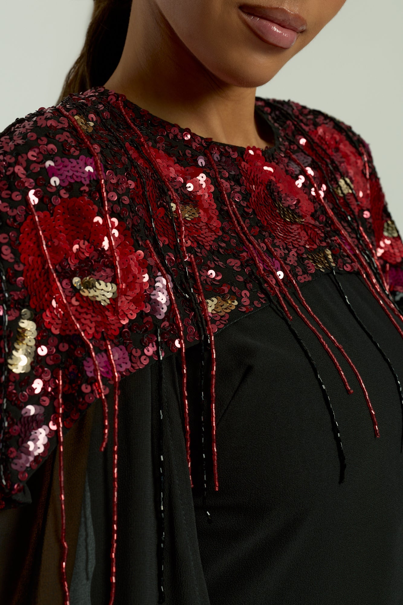 Elegant black maxi cape dress with a relaxed, fluid silhouette and a sheer flowing cape overlay that offers arm and shoulder coverage. The cape is decorated with deep red hand-sequinned floral motifs and cascading beaded tassels that add movement and spar