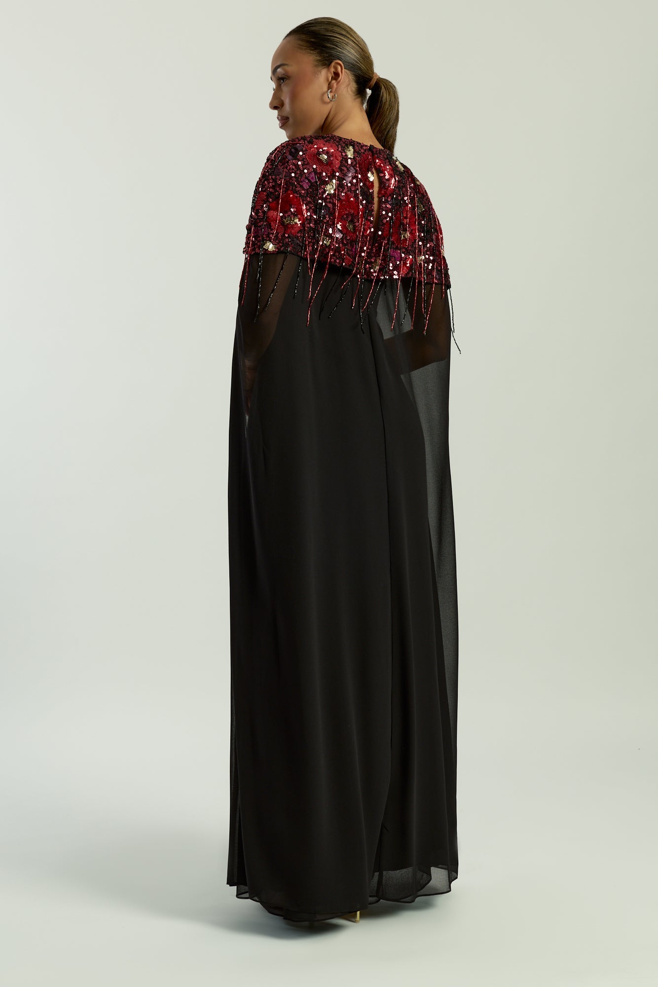 Elegant black maxi cape dress with a relaxed, fluid silhouette and a sheer flowing cape overlay that offers arm and shoulder coverage. The cape is decorated with deep red hand-sequinned floral motifs and cascading beaded tassels that add movement and spar