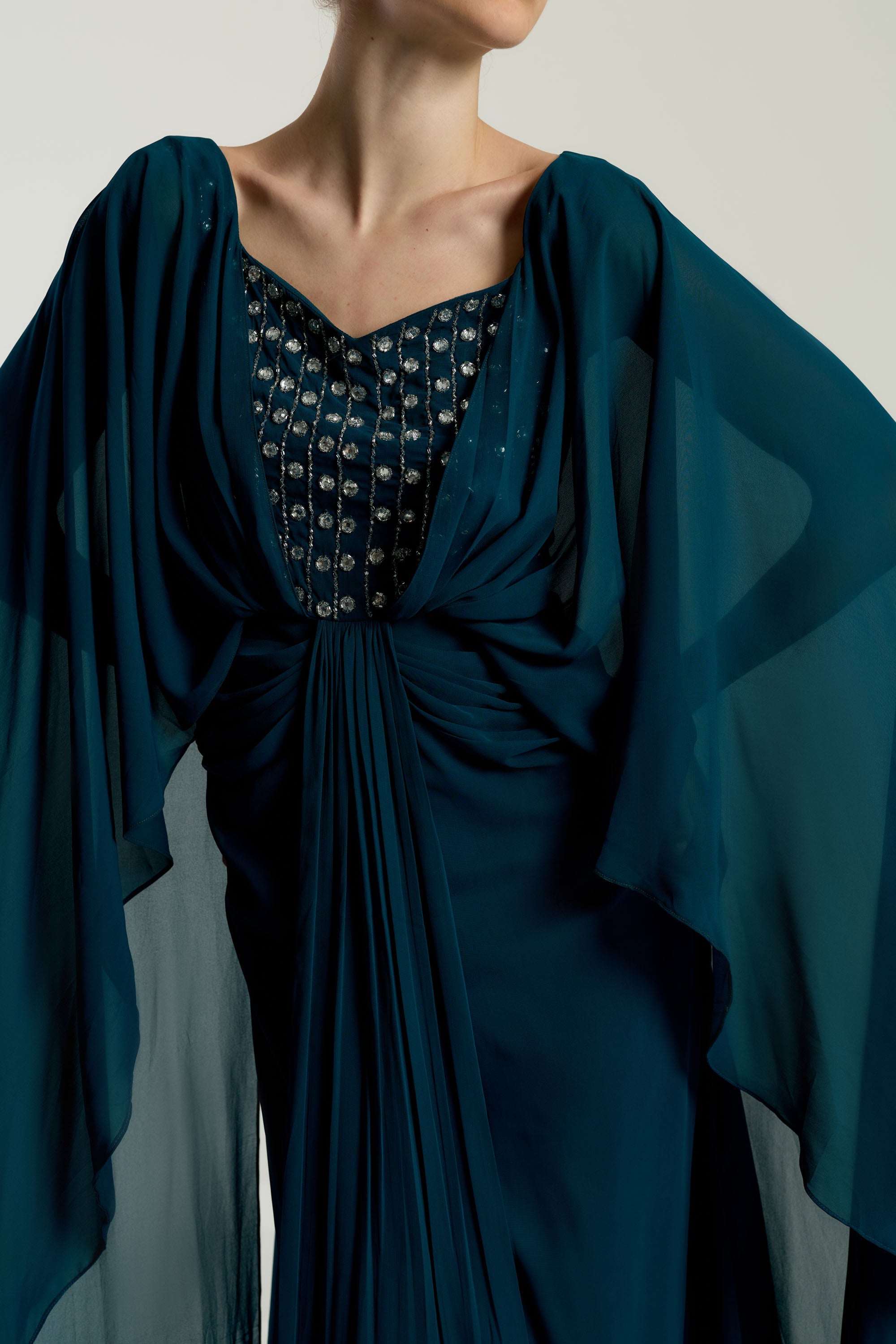 Rhea teal cape sleeve maxi dress with silver-embellished bodice, flowing cape overlay, and modest elegant silhouette for weddings and formal events.