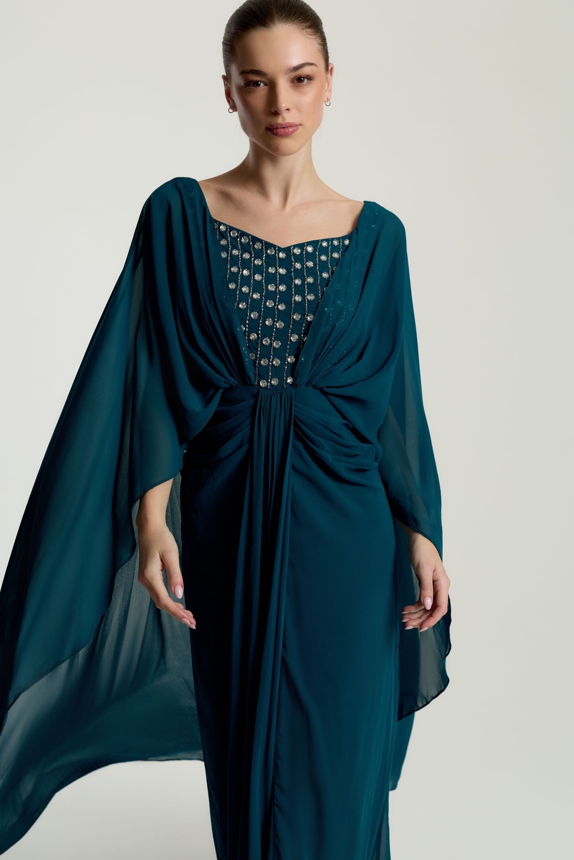 Rhea teal cape sleeve maxi dress with silver-embellished bodice, flowing cape overlay, and modest elegant silhouette for weddings and formal events.
