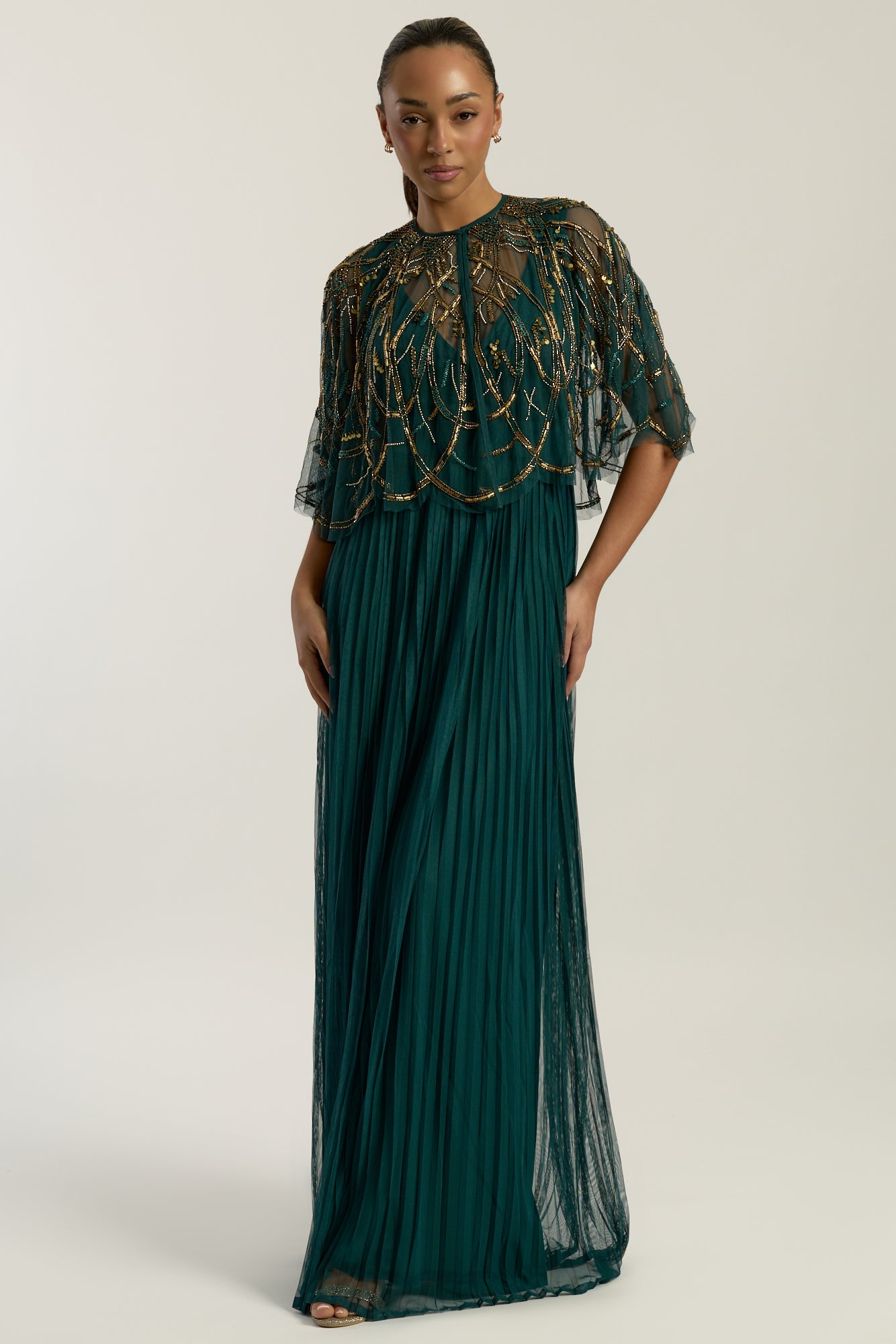Green embellished maxi dress with flowing pleated skirt and detachable sheer cape overlay, featuring gold bead detailing and an elegant V-neck silhouette for occasion wear.
