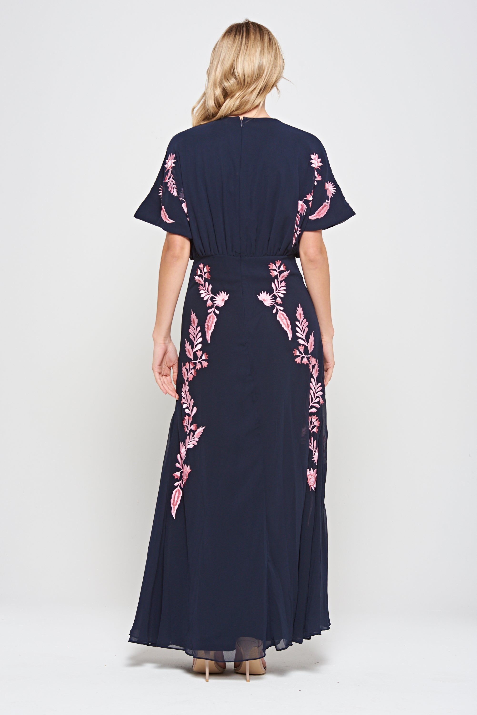 Navy floral embroidered maxi dress with pink detailing, worn by a model. Features flutter sleeves, a fitted waist and a flowing full-length skirt—perfect for weddings, evening occasions and special events.