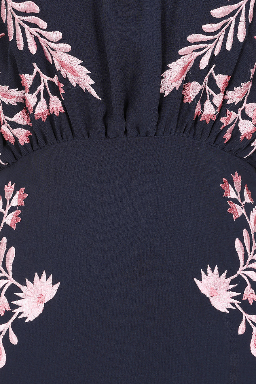 Navy floral embroidered maxi dress with pink detailing, worn by a model. Features flutter sleeves, a fitted waist and a flowing full-length skirt—perfect for weddings, evening occasions and special events.
