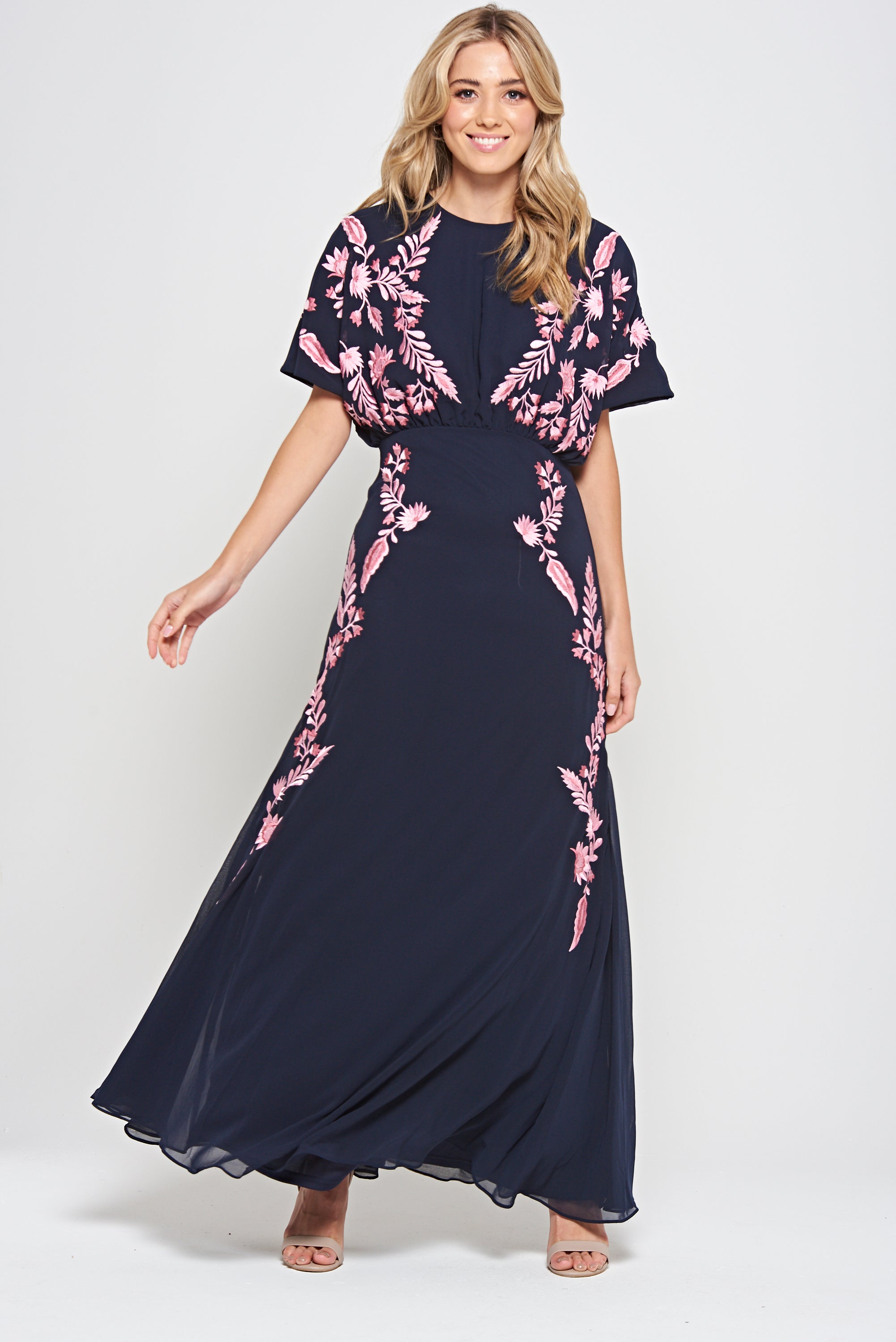 Navy floral embroidered maxi dress with pink detailing, worn by a model. Features flutter sleeves, a fitted waist and a flowing full-length skirt—perfect for weddings, evening occasions and special events.